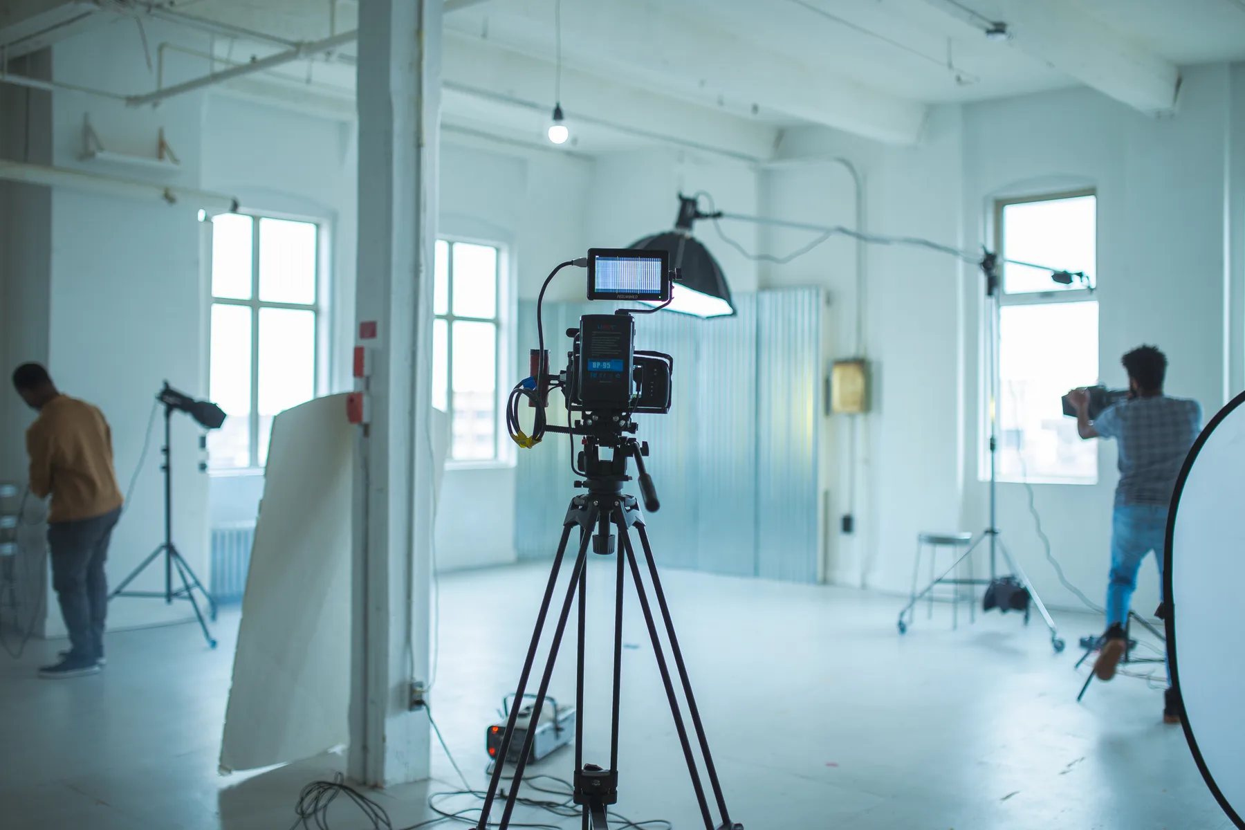 A camera on a tripod in a bright studio, with two of the crew setting up lights behind it